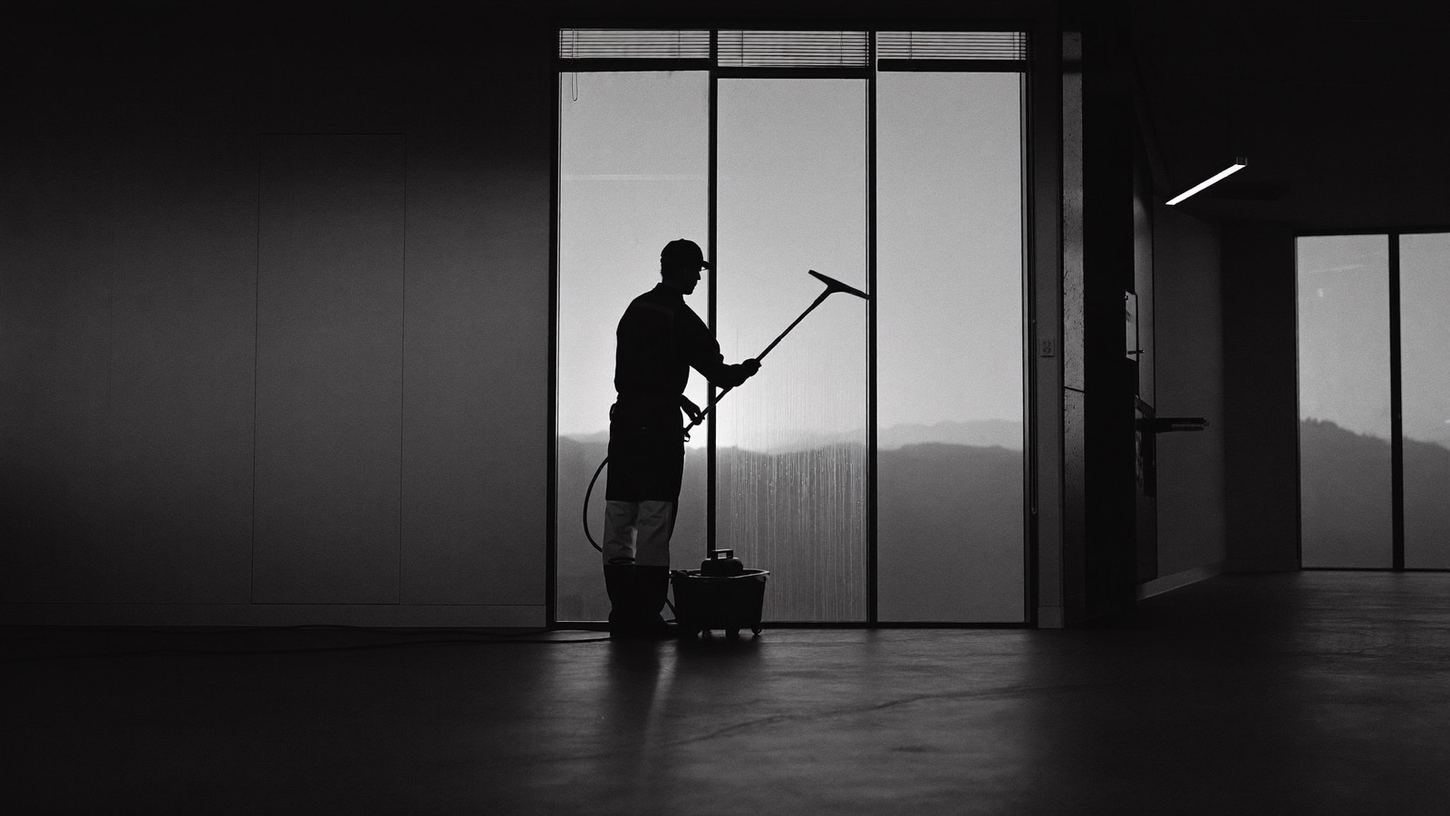 A commercial cleaner squeegeeing a tall office window at dawn, silhouetted against the light
